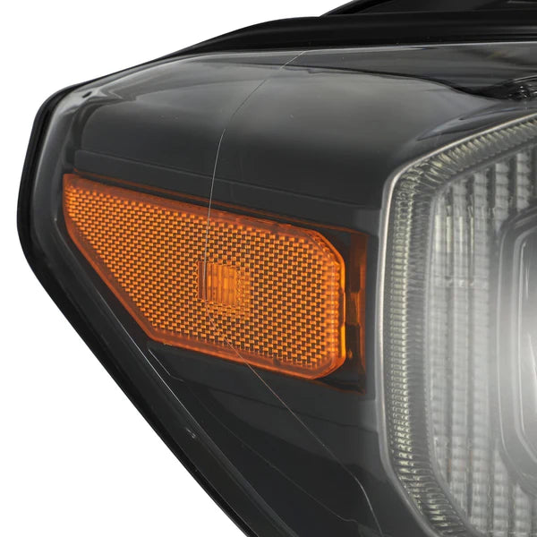 Close-up view of a Toyota Tacoma's headlight assembly, showcasing the intricate design of the orange turn signal light and the surrounding black and clear components, featuring AlphaRex USA NOVA-Series LED Projector Headlights in Mid-Night Black for enhanced visibility (SKU: 880705), compatible with 2016-2023 models.