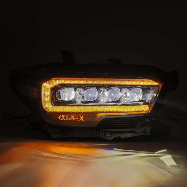 A close-up of the AlphaRex NOVA-Series LED Projector Headlight - Mid-Night Black - 2016-2023 Toyota Tacoma (SKU: 880705) by AlphaRex USA, illuminated against a dark background. The headlight showcases a distinctive outline of amber LED lights surrounding multiple bright white LED beams. A subtle orange light reflects on the surface below the headlight.