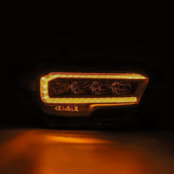 Close-up of a Toyota Tacoma headlight in the dark, illuminated with amber LED lights. The AlphaRex USA AlphaRex NOVA-Series LED Projector Headlights - Mid-Night Black (SKU: 880705) for 2016-2023 Toyota Tacoma boasts a modern, rectangular design with several circular light elements inside, and a slight glow emanates from the surrounding area.