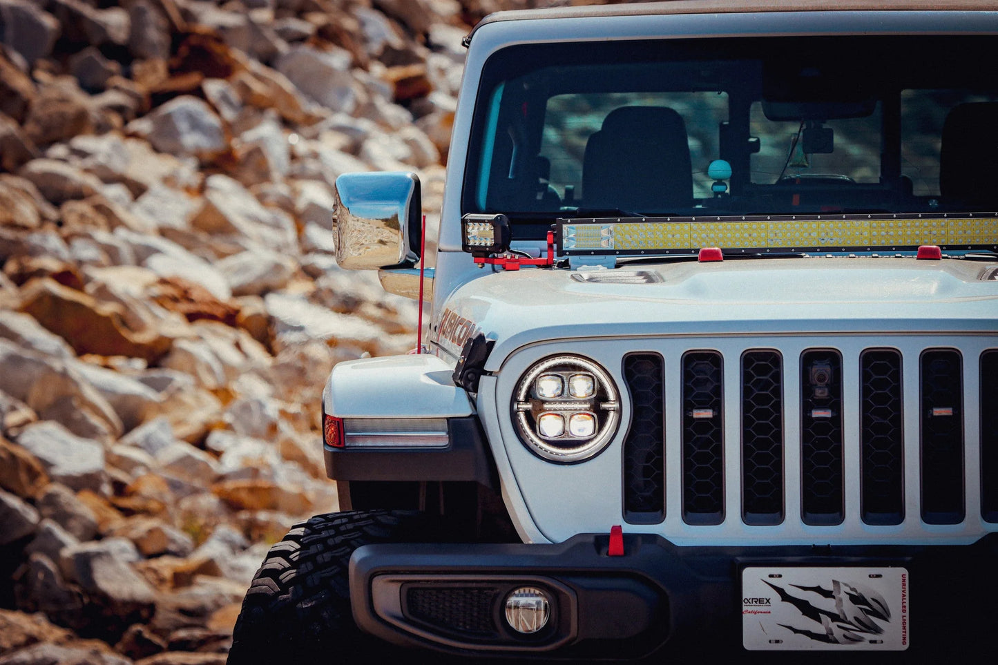 A white Jeep Wrangler JL off-road vehicle, featuring rugged tires and a prominent front grille with distinct circular AlphaRex USA NOVA-Series LED Projector Headlights in black (SKU: 880868), is parked near rocky terrain. The vehicle is equipped with additional lighting and a visible custom license plate.