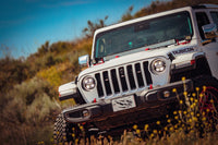 A white Jeep Rubicon navigating off-road terrain, surrounded by shrubs and wildflowers under a clear blue sky. The vehicle is equipped with large tires and AlphaRex USA AlphaRex NOVA-Series LED Projector Headlights in black for the 2018-2023 Jeep Wrangler JL/Gladiator JT (SKU: 880868), showcasing its rugged design and off-road capabilities.