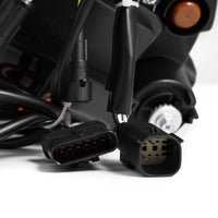 A close-up image of two black electrical connectors with multiple pins, attached to a bundle of wires. The connectors are prominently positioned in the foreground against a blurred background featuring various mechanical and electrical components, including AlphaRex USA's AlphaRex NOVA-Series LED Projector Headlights for 2018-2023 Jeep Wrangler JL/Gladiator JT (SKU: 880868).