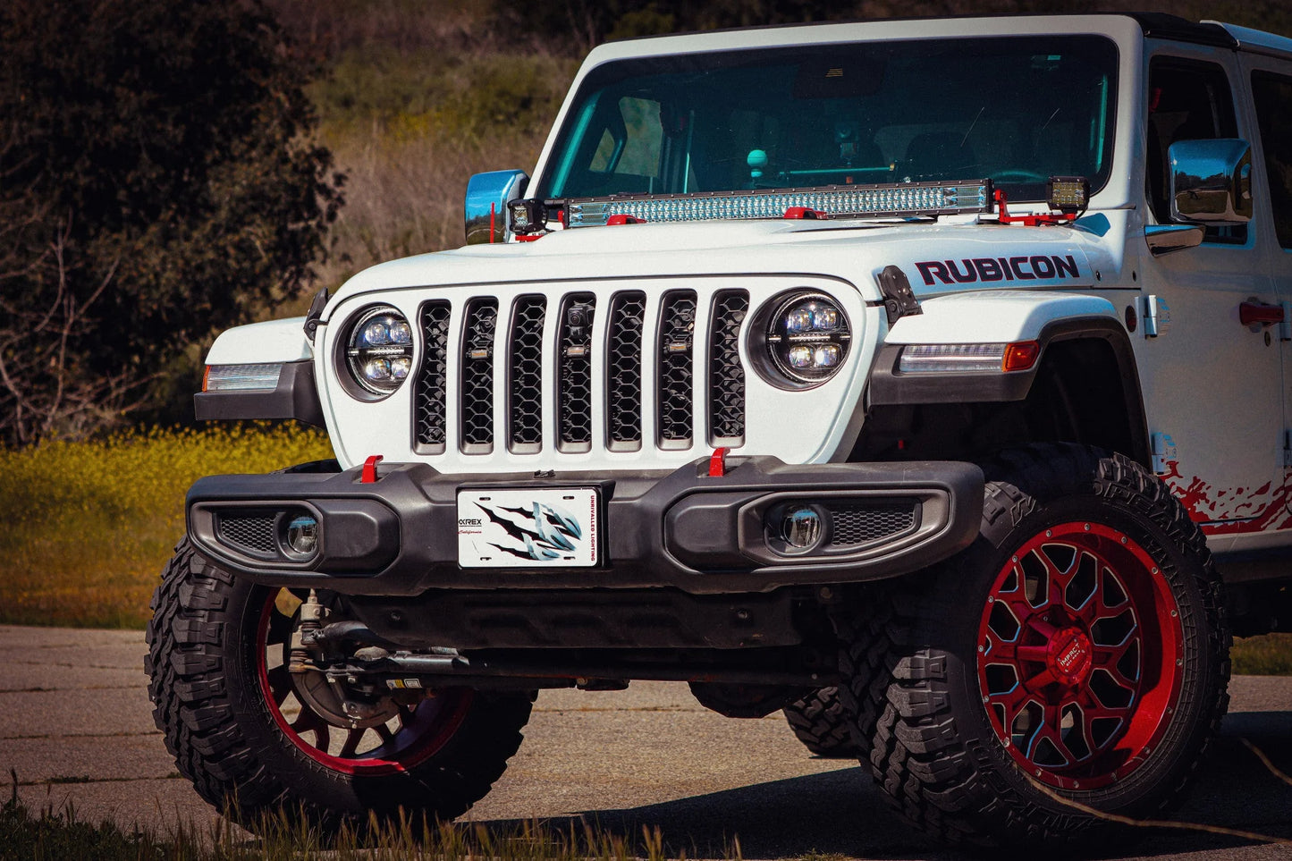 A white Jeep Wrangler JL with "Rubicon" on the hood, featuring custom red rims and large tires, is parked on a paved road against a grassy backdrop. The vehicle boasts AlphaRex USA's NOVA-Series LED Projector Headlights in black (SKU: 880868) for the 2018-2023 Jeep Wrangler JL/Gladiator JT, along with a rugged front bumper designed for robust off-road performance.