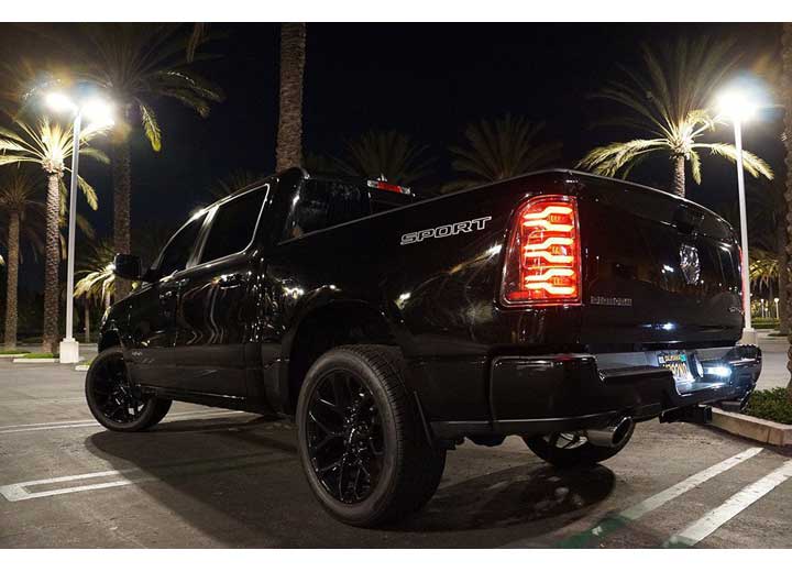 A black pickup truck with the word "SPORT" on its side is parked in a well-lit area at night, surrounded by tall palm trees. The truck's taillights are on, featuring AlphaRex USA's RAM LUXX LED Tail Lights Alpha-Black with Sequential Signal Lights for the 19-22 model, enhancing visibility. It also boasts black wheels and tinted windows.