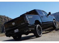 A black Dodge RAM 1500 pickup truck is shown from the rear at an upward angle, parked on a rocky, off-road terrain with mountains in the background. Its "4x4 Off Road" badge is visible on the side, and the truck features custom wheels and an aftermarket exhaust. Additionally, it boasts AlphaRex USA's RAM LUXX LED Tail Lights Alpha-Black with Sequential Signal Lights for enhanced visibility.