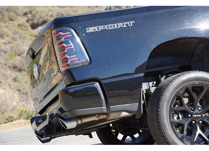 Close-up of the rear end of a black pickup truck with the word "SPORT" written on the side. The image focuses on the RAM LUXX LED Tail Lights Alpha-Black with Sequential Signal Lights - 19-22 Model, dual exhaust pipes, and black alloy wheels by AlphaRex USA. The background features a hilly landscape with sparse vegetation.