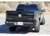 A black Dodge Ram 1500 Big Horn 4x4 truck, featuring RAM LUXX LED Tail Lights in Alpha-Black with Sequential Signal Lights by AlphaRex USA, is parked on a paved area with mountains in the background. The tailgate and exhaust pipes are visible, along with a license plate labeled "OREX." The truck has tinted windows and large tires.