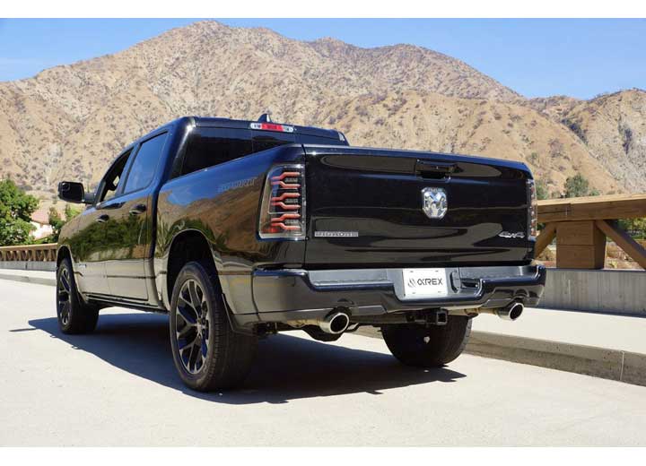 A black Ram pickup truck is parked on a road with mountainous terrain in the background. The vehicle boasts enhanced visibility thanks to RAM LUXX LED Tail Lights Alpha-Black with Sequential Signal Lights by AlphaRex USA for 19-22 models, along with tinted rear lights, dual exhaust pipes, and a "6'4" badge visible on its bed.