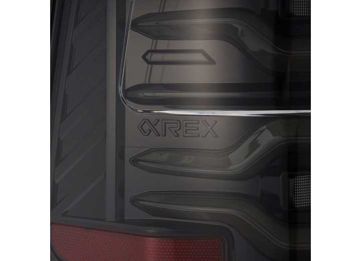 Close-up of the rear end of a dark-colored vehicle, showcasing a futuristic design with the word "OREX" prominently displayed in the center. Visible are sleek, angular elements and part of AlphaRex USA's RAM LUXX LED Tail Lights Alpha-Black with Sequential Signal Lights for 19-22 models at the bottom left corner, offering enhanced visibility with sequential signal lights.