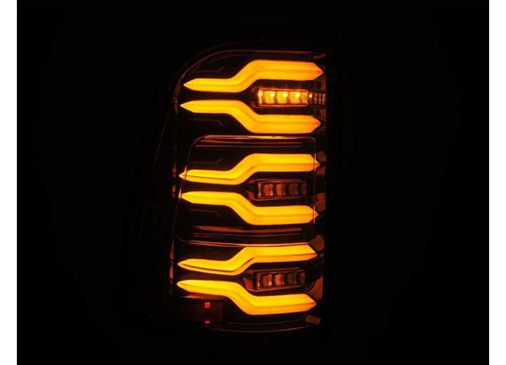 Close-up of a vehicle's illuminated RAM LUXX LED Tail Lights Alpha-Black by AlphaRex USA, showcasing a unique design with sequential signal lights and multiple horizontal, curved, amber-colored LED light strips arranged vertically against a black background. Enhanced visibility ensures safety and style. Suitable for 19-22 models.