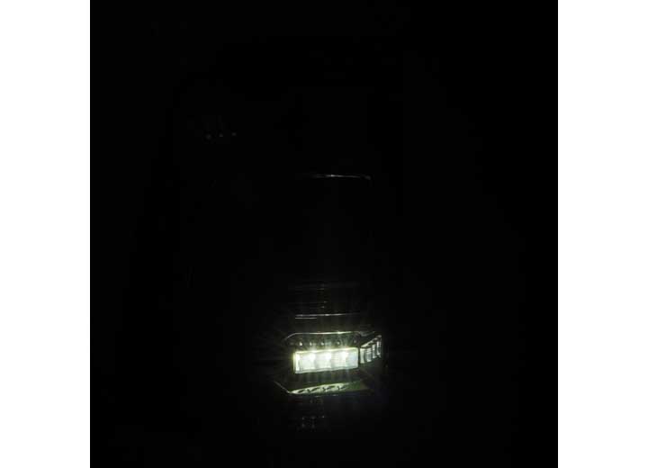 A close-up shot of a bright, white LED light in the darkness, partially illuminating what appears to be part of a vehicle's headlight assembly. The RAM LUXX LED Tail Lights Alpha-Black with Sequential Signal Lights - 19-22 Model by AlphaRex USA provide enhanced visibility as the primary source of light in the otherwise pitch-black surroundings.