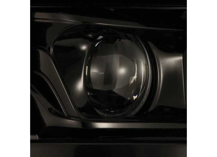 Close-up photograph of the 21-23 F150 LUXX LED Projector Headlights Alpha-Black with ACT Light & Sequential Signal by AlphaRex USA. The headlight lens reflects light, showcasing intricate details of the headlight housing. The image focuses on the circular shape of the headlight in a glossy black finish.