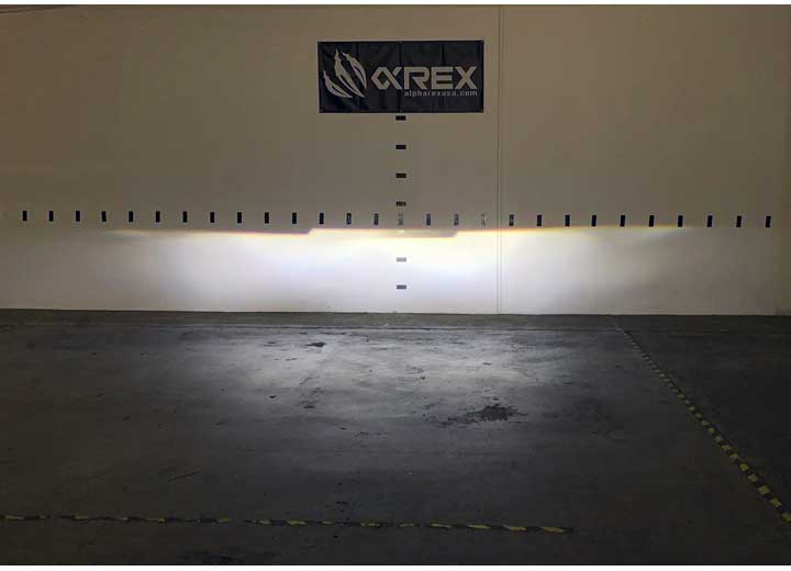 A warehouse wall with a black banner featuring the word "AlphaRex USA" and a logo, illuminated by a bright horizontal strip of light. The concrete floor and lower wall are dimly lit, with yellow caution tape markings on the floor. The space highlights the 09-18 RAM 2500 LED Projector Headlights w/ ACT Light / Seq Signal and DRL Alpha.