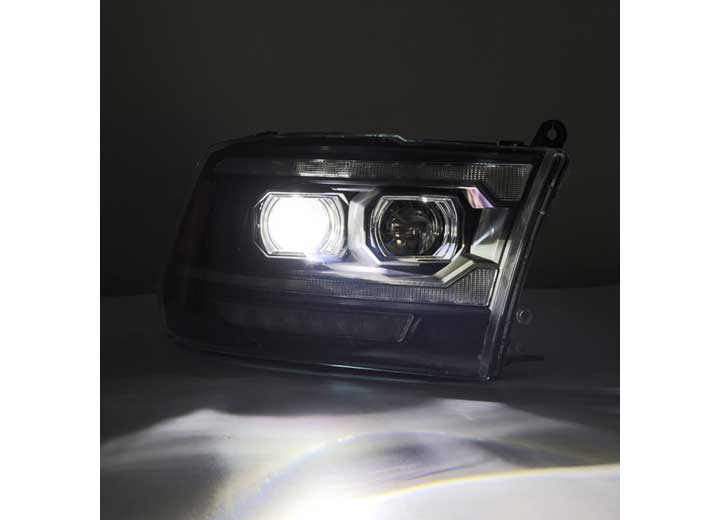 A close-up of a car's headlight assembly, featuring the AlphaRex USA 09-18 RAM 2500 LED Projector Headlights with ACT Light, Sequential Signal, and DRL Alpha, shows one illuminated light emitting a bright white beam. The headlight boasts a modern, sleek design with multiple reflective surfaces and clear glass. The dark background accentuates the brightness of the light.