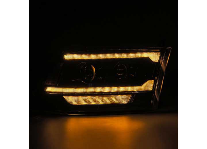 Close-up image of the 09-18 RAM 2500 LED Projector Headlights with ACT Light, Sequential Signal, and DRL Alpha by AlphaRex USA in the dark. The headlights showcase a modern design with multiple horizontal light strips emitting a bright amber glow. The sleek lines and illuminated elements suggest a futuristic automotive design.