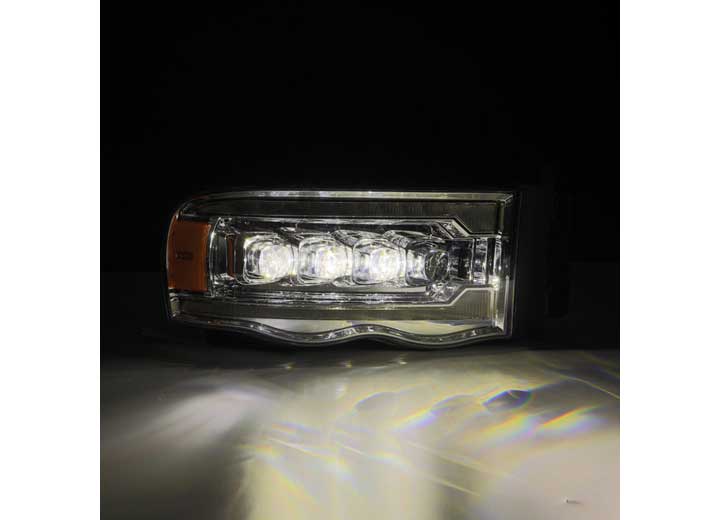 Close-up of a modern AlphaRex USA RAM 1500/2500/3500 2002-2005/2003-2005 Nova-Series LED Projector Headlight - Alpha Black turned on, emitting bright white light. The headlight has a sleek design with multiple LEDs visible inside and a small amber-colored section on the left. The dark background enhances visibility from the Alpha Black headlight's illumination.