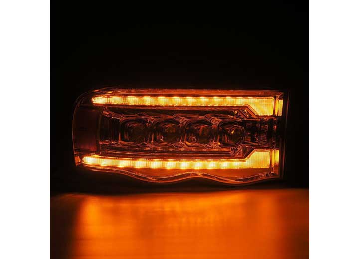 A brightly lit amber LED tail light with multiple light segments is illuminated against a dark background, casting a yellowish-orange glow on the surface below. Complemented by the AlphaRex USA RAM 1500/2500/3500 2002-2005/2003-2005 Nova-Series LED Projector Headlights in Alpha Black, it ensures enhanced visibility as part of the vehicle's lighting system.