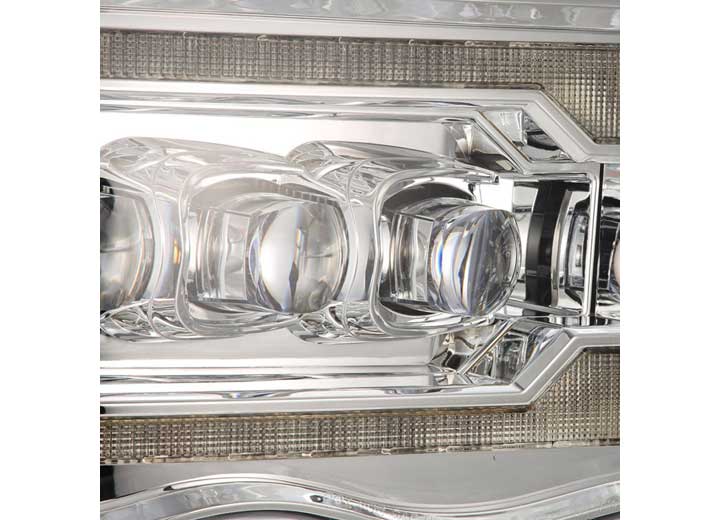 Close-up image of an AlphaRex USA Alpha Black headlight on a RAM 1500/2500/3500 2002-2005/2003-2005 Nova-Series LED Projector Headlight. The headlight features a sleek and intricate design with multiple reflective and transparent elements, including geometric shapes and a textured background. The metallic and glass components create a futuristic and detailed appearance, ensuring enhanced visibility.