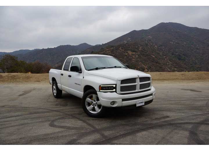 A white pickup truck with a quad cab is parked on a wide asphalt area in front of a scenic mountainous backdrop under a cloudy sky. The license plate reads "C. JOHNSON." The truck's AlphaRex USA RAM 1500/2500/3500 2002-2005/2003-2005 Nova-Series LED Projector Headlights in Alpha Black offer enhanced visibility, showcasing its front grille and headlights.