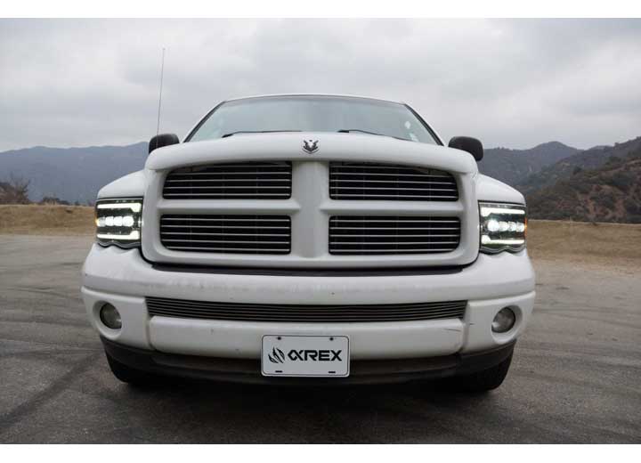 Front view of a white Dodge Ram truck with a custom grille and AlphaRex USA RAM 1500/2500/3500 2002-2005/2003-2005 Nova-Series LED Projector Headlights in Alpha Black for enhanced visibility, parked on a road with mountains and a cloudy sky in the background. The truck has a "QREX" logo on the front license plate.