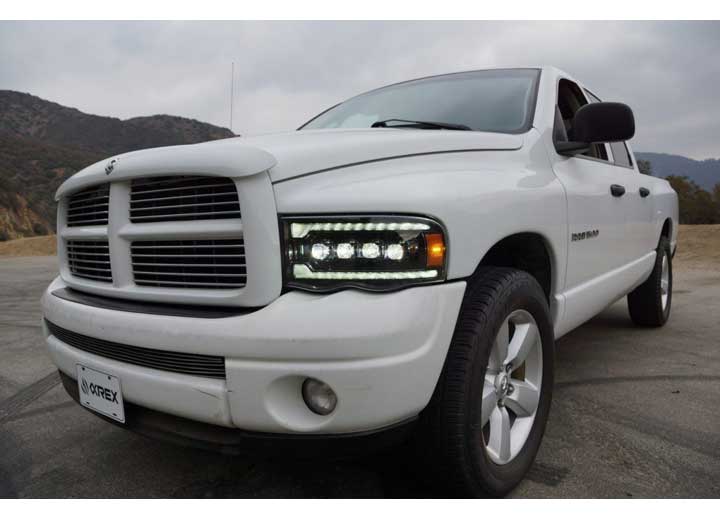 A white Dodge Ram pickup truck is parked on a road with mountains in the background. The truck's enhanced visibility is showcased by its AlphaRex USA RAM 1500/2500/3500 2002-2005/2003-2005 Nova-Series LED Projector Headlights in Alpha Black, which are turned on, emphasizing the prominent front grille and sporty design. The image is taken from a low angle, highlighting the vehicle’s front and side.