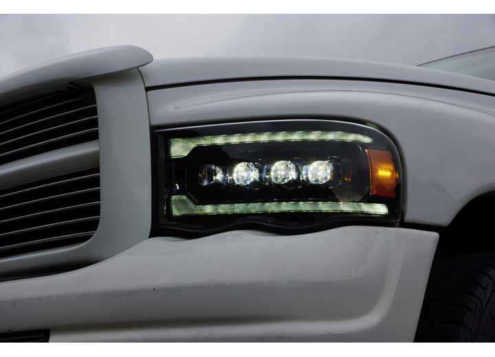 Close-up of the front left corner of a white truck showcasing its AlphaRex USA RAM 1500/2500/3500 2002-2005/2003-2005 Nova-Series LED Projector Headlights in Alpha Black with multiple lights for enhanced visibility. The vehicle grill and part of the hood are also visible against an overcast sky.