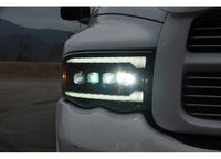 Close-up view of the front left headlight of a white truck illuminated at dusk, featuring AlphaRex USA RAM 1500/2500/3500 2002-2005/2003-2005 Nova-Series LED Projector Headlights - Alpha Black for enhanced visibility. The background shows a blurred outdoor setting with a mountain and cloudy sky.