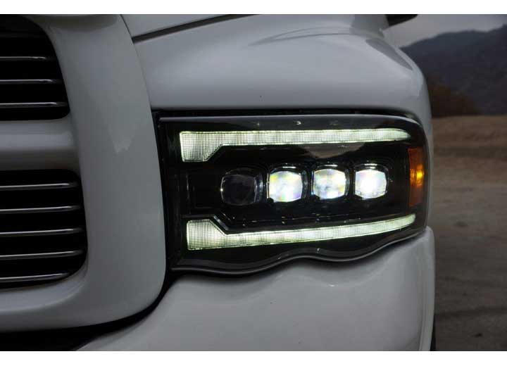 Close-up of the front left headlight of a modern white vehicle, featuring AlphaRex USA RAM 1500/2500/3500 2002-2005/2003-2005 Nova-Series LED Projector Headlights in Alpha Black. The headlights have a sleek, horizontal design with multiple LEDs enclosed in a rectangular housing. The vehicle's grille is partially visible to the left. The background is blurry.