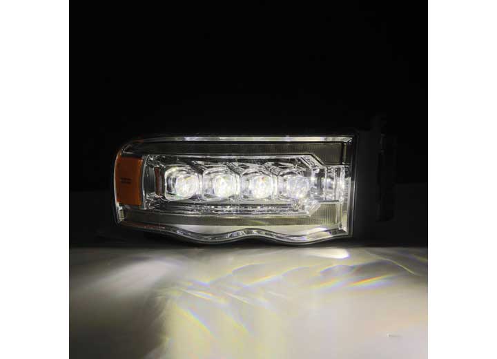 Close-up of a vehicle headlight with a black background. The AlphaRex USA RAM 1500/2500/3500 2002-2005/2003-2005 Nova-Series LED Projector Headlights in Alpha Black are illuminated, showcasing multiple LED lights within a rectangular housing that features a clear lens and an amber section on the left side. Reflections are visible on the surface below, enhancing visibility.
