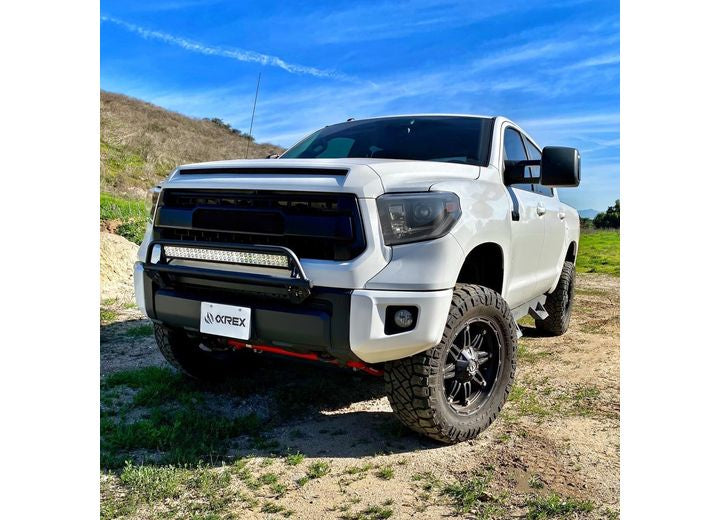 A white pickup truck with lifted suspension and large off-road tires is parked on a dirt path outdoors. The truck, featuring a rugged front grille, mounted light bar, black rims, and AlphaRex USA's Tundra 14-21 LED Projector Headlights with Sequential Signal & DRL, also boasts LED headlights. The sky is clear and blue with a grassy hill in the background.