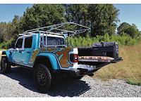 A customized blue and orange 2020 Jeep Gladiator, equipped with a roof rack and the CUSTOM ORDER CLBEDSLIDE 1000 SILVER CLASSIC 57IN X 43IN from Bedslide, is parked on gravel near a grassy area with trees in the background. The bed slide is extended, revealing additional cargo space and gear.