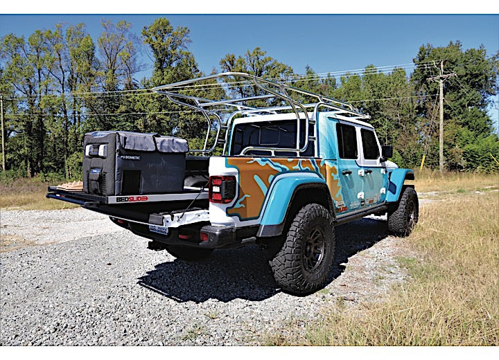 A modified blue Jeep Gladiator pickup truck with a camo pattern on its rear sides, featuring a large roof rack and an attached cargo carrier at the back, is parked on a gravel path. Trees and vegetation are visible in the background. The truck also boasts the Bedslide CUSTOM ORDER CLBEDSLIDE 1000 SILVER CLASSIC 57IN X 43IN for easy access to bed contents.