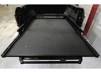 A truck bed with a Bedslide BEDSLIDE 1000 CLASSIC BLACK 68IN X 48IN sliding platform installed is extended outward, covering most of the truck bed space. The surrounding area appears to be a garage or workshop with a concrete floor and a red vertical beam to the right.