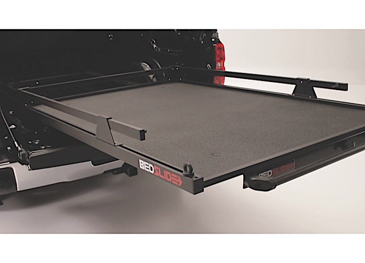 A 73-inch by 48-inch BEDSLIDE MAX EXTENSION 1500 in black, from the Bedslide brand, is partially extended from the back of a black truck. This flat-surfaced bedslide extension with raised sides is designed for easy loading and unloading of goods. The background is plain and light-colored.