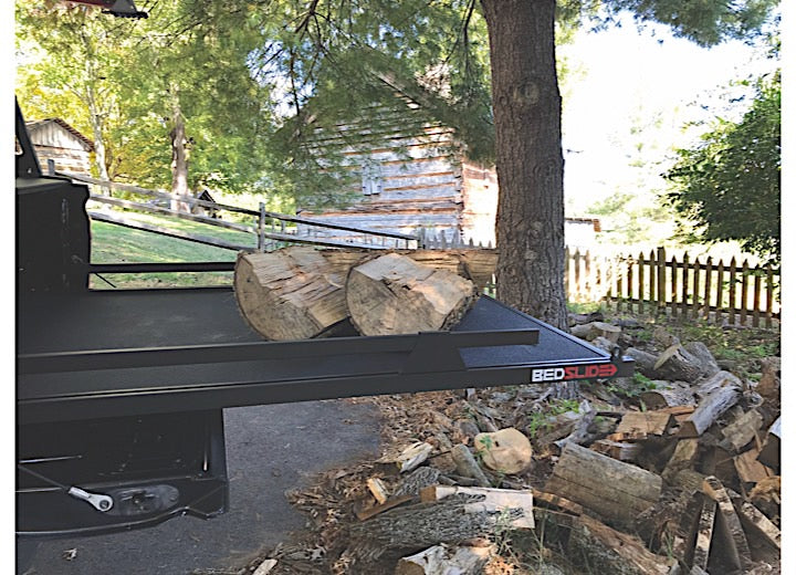 A truck bed equipped with the Bedslide 73IN x 48IN BEDSLIDE MAX EXTENSION 1500 in black, loaded with chopped firewood, is parked on a paved driveway. Additional firewood is scattered on the ground nearby. In the background, trees and a rustic wooden building enclosed by a wooden fence can be seen.