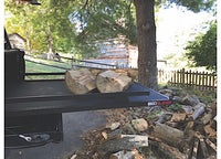 A truck bed equipped with the Bedslide 73IN x 48IN BEDSLIDE MAX EXTENSION 1500 in black, loaded with chopped firewood, is parked on a paved driveway. Additional firewood is scattered on the ground nearby. In the background, trees and a rustic wooden building enclosed by a wooden fence can be seen.