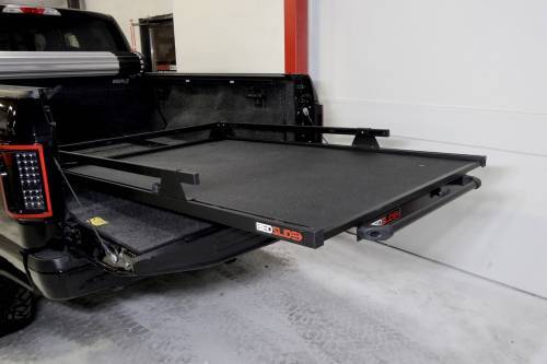 The BEDSLIDE 1000 CLASSIC BLACK 63IN X 47IN, a truck bed slide from Bedslide, is partially extended from the back of a black pickup truck. Featuring a sturdy metal frame and flat surface, it significantly enhances the truck bed's functionality for easier loading and unloading of heavy loads. The pickup is parked in a garage with a concrete floor.