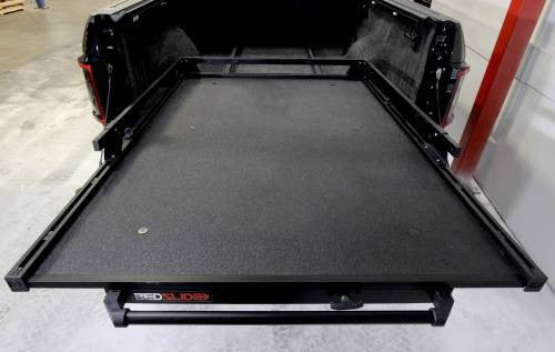 A black truck bed fitted with the BEDSLIDE 1000 CLASSIC BLACK 63IN X 47IN underscores its exceptional truck bed functionality. This slide, built to handle heavy loads, boasts a textured surface and prominently showcases the Bedslide logo near the tailgate. The truck is parked in a well-lit garage with visible concrete flooring and shelving.