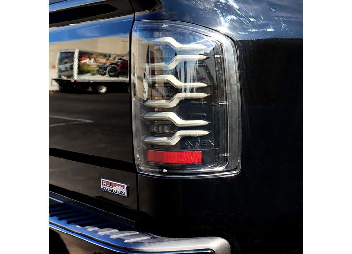 Close-up of a 14-18 Sierra 1500/2500/3500HD exhibiting AlphaRex USA LUXX LED Tail Lights in Alpha-Black, featuring distinctive wave-like white lines and a red section at the bottom. The vehicle's black exterior includes an "All Terrain" sticker near the tail light, set against an outdoor parking area backdrop.