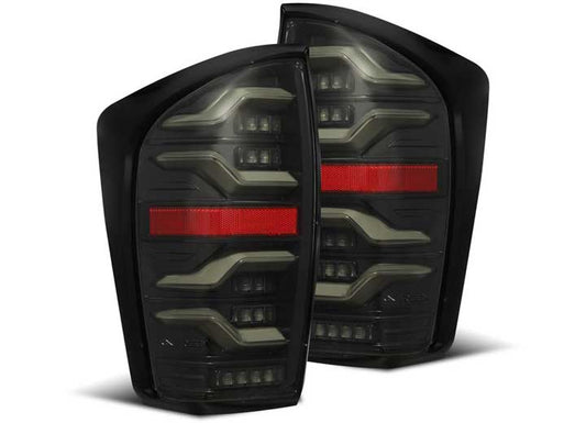 A pair of AlphaRex USA Tacoma 16-23 Luxx-Series LED Tail Lights in an Alpha-Black finish, featuring clear lenses with intricate internal detailing and a horizontal red strip in the middle. These black LED taillights also include sequential amber signals for enhanced visibility, positioned upright and slightly angled to display their design features.