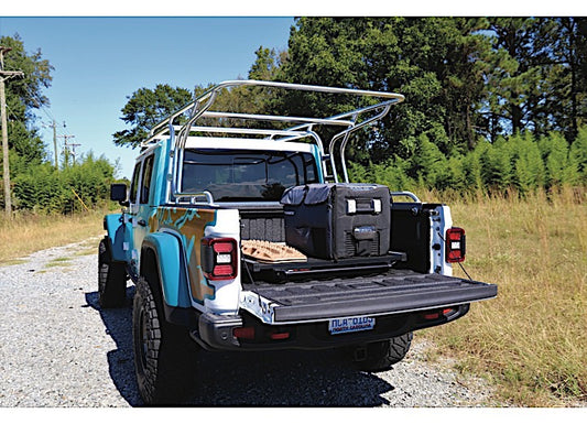 A teal pickup truck with a silver metal rack installed on top is parked on a gravel path. The truck's tailgate is down, revealing a black storage container and a beige-colored item on the Bedslide CUSTOM ORDER CLBEDSLIDE 1000 SILVER CLASSIC 57IN X 43IN bed slide in the truck bed of a 2020 Jeep Gladiator. The background includes grass and trees under a clear sky.