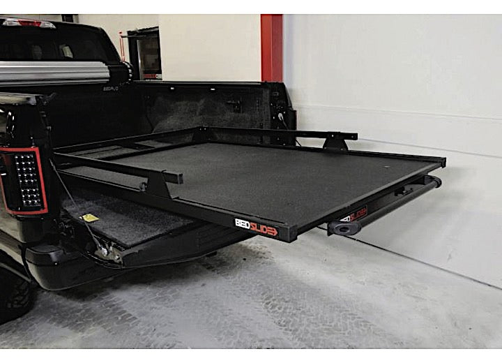 A truck with its tailgate open reveals a Bedslide BEDSLIDE 1000 CLASSIC BLACK 68IN X 48IN, an industrial-grade sliding platform that makes loading and unloading effortless. Parked indoors next to white and red walls, this setup highlights both functionality and rugged aesthetics.