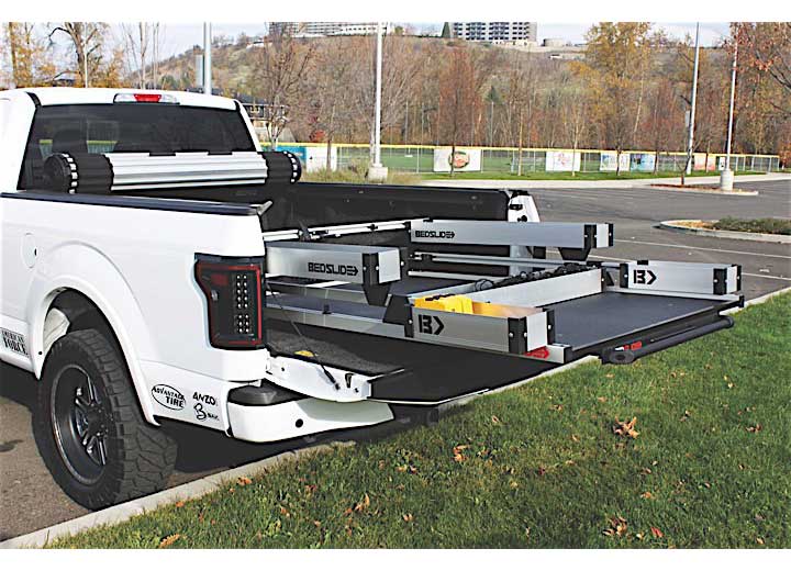 A white pickup truck with an open tailgate parked outdoors features the Bedslide COMPLETE BEDBIN KIT (BSA-DK, MK, SK) with multiple sliding trays and compartments, maximizing storage space and showcasing organized solutions. The scene is framed by trees with autumn leaves, hinting at the system's easy installation for ultimate bed storage.