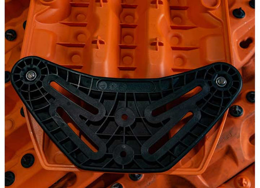 Close-up of an orange plastic surface with a mounted MAXTRAX FLAT RACK MOUNT WITH T-BOLT AND XTREME MPS 17MM attached by two screws. The black component features several elongated slots and geometric patterns, contrasting starkly against the bright background, resembling the rugged design of an off-road accessory such as recovery tracks by MAXTRAX.